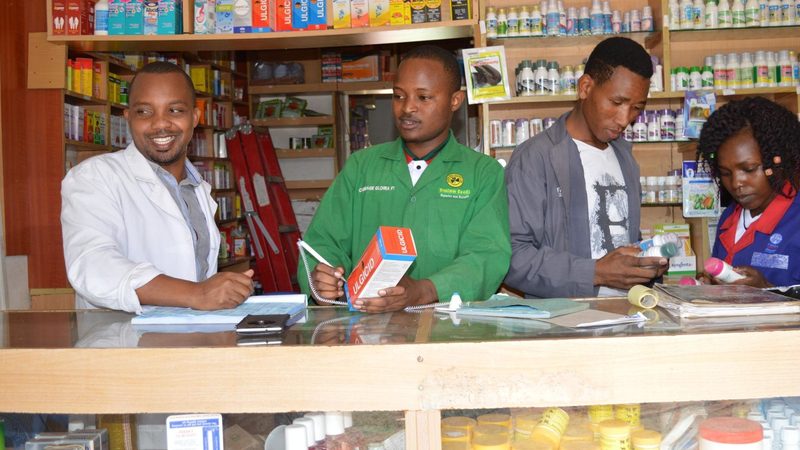 Pharmacy staff serving at the counter, one writing in a paper record book