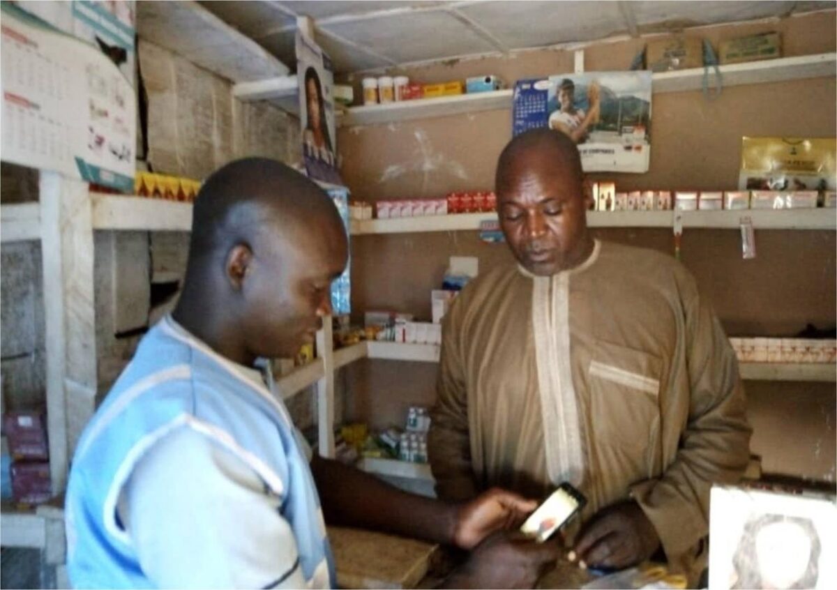 A shopkeeper checking information on a mobile phone with a colleague inside a small shop